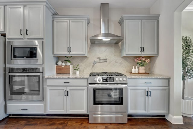 kitchen featuring wall chimney range hood, light stone counters, tasteful backsplash, stainless steel appliances, and dark wood-type flooring