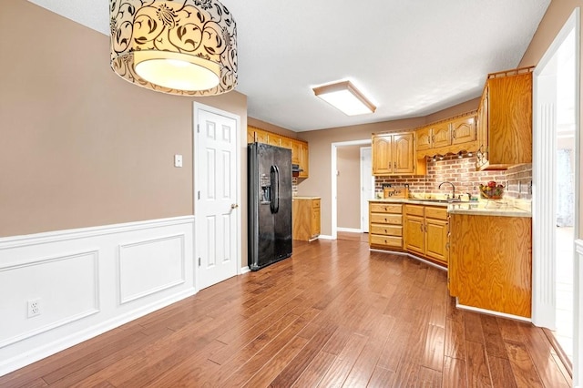 kitchen with a sink, dark wood-style flooring, black refrigerator with ice dispenser, light countertops, and wainscoting