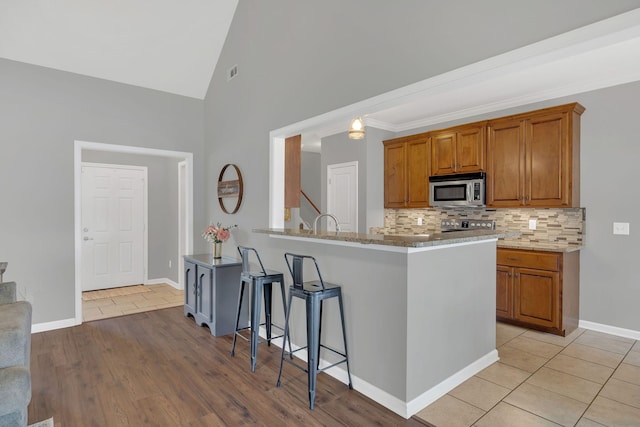 kitchen featuring light stone counters, brown cabinetry, stainless steel microwave, baseboards, and backsplash