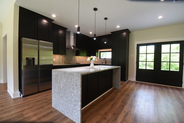 kitchen with a kitchen island, backsplash, dark cabinets, stainless steel fridge, and wall chimney exhaust hood