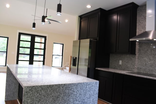 kitchen featuring wall chimney range hood, decorative backsplash, light stone countertops, and stainless steel fridge