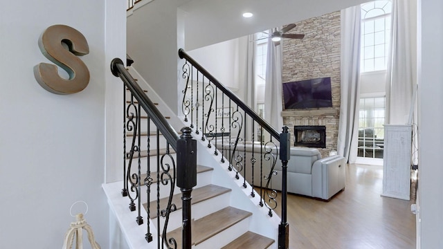staircase featuring wood finished floors, ceiling fan, a high ceiling, and a fireplace