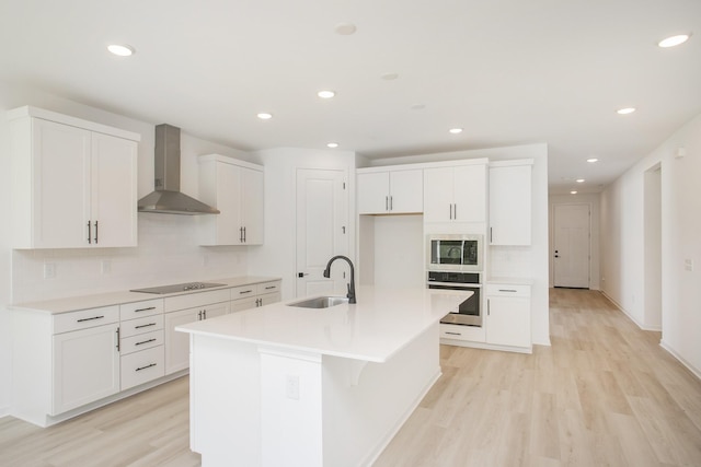kitchen featuring wall chimney range hood, appliances with stainless steel finishes, a sink, light wood finished floors, and tasteful backsplash