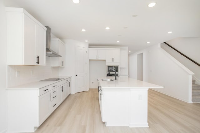 kitchen with a sink, black electric cooktop, light wood-style flooring, wall chimney exhaust hood, and tasteful backsplash