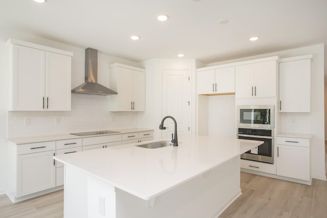 kitchen with an island with sink, wall chimney range hood, a sink, light wood-style flooring, and stainless steel appliances