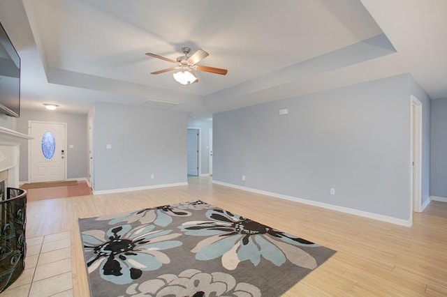 unfurnished living room featuring ceiling fan, a tiled fireplace, baseboards, a tray ceiling, and light wood-style flooring