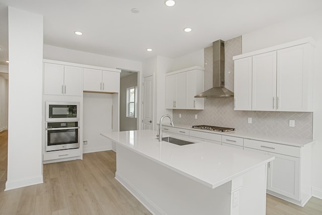 kitchen featuring wall chimney range hood, tasteful backsplash, a sink, light wood finished floors, and appliances with stainless steel finishes