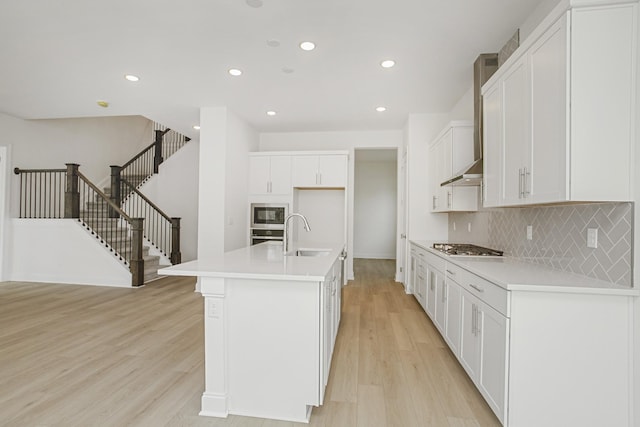 kitchen featuring wall chimney range hood, tasteful backsplash, light wood-type flooring, a sink, and stainless steel appliances
