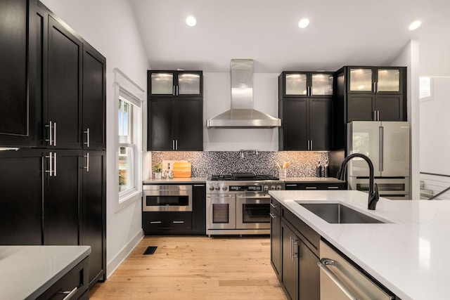 kitchen with wall chimney exhaust hood, tasteful backsplash, dark cabinetry, a sink, and appliances with stainless steel finishes
