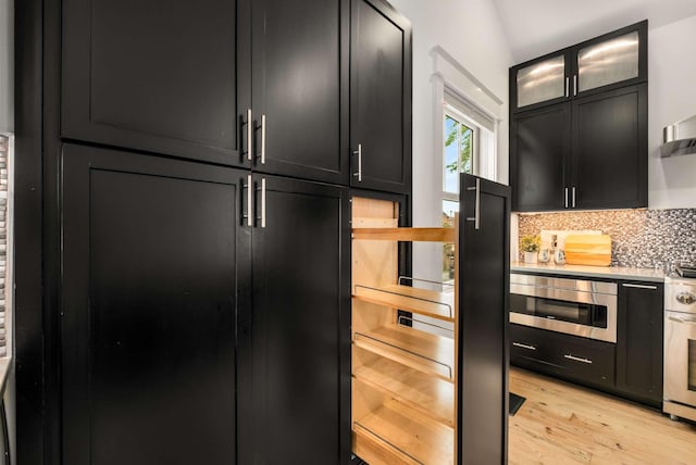 kitchen with decorative backsplash, glass insert cabinets, stainless steel microwave, dark cabinets, and light wood finished floors