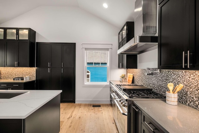 kitchen featuring lofted ceiling, wall chimney exhaust hood, dark cabinetry, backsplash, and double oven range