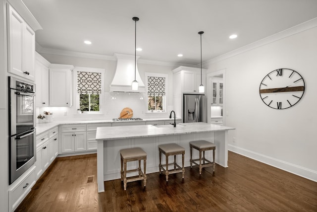 kitchen with white cabinets, premium range hood, stainless steel appliances, backsplash, and a center island with sink
