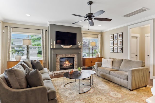 living room featuring a brick fireplace, ceiling fan, visible vents, and crown molding