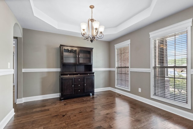 unfurnished dining area with a raised ceiling, dark wood finished floors, a chandelier, and baseboards