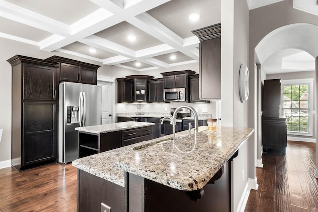 kitchen with stainless steel appliances, arched walkways, a kitchen island, a sink, and dark brown cabinetry