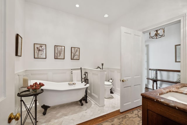 full bathroom featuring a freestanding bath, vanity, a decorative wall, toilet, and a wainscoted wall