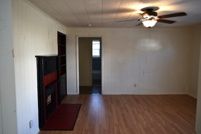 unfurnished living room featuring a fireplace with flush hearth, ceiling fan, wooden walls, and wood finished floors