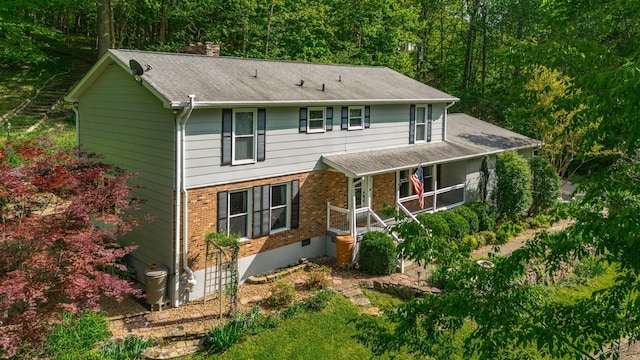 traditional-style house featuring a shingled roof, brick siding, a chimney, a forest view, and covered porch