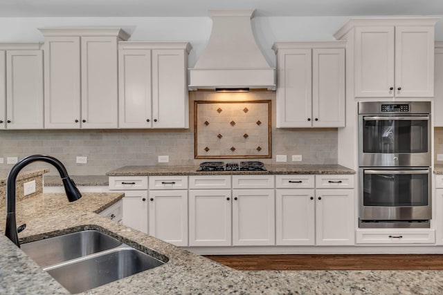 kitchen featuring light stone counters, a sink, double oven, premium range hood, and backsplash