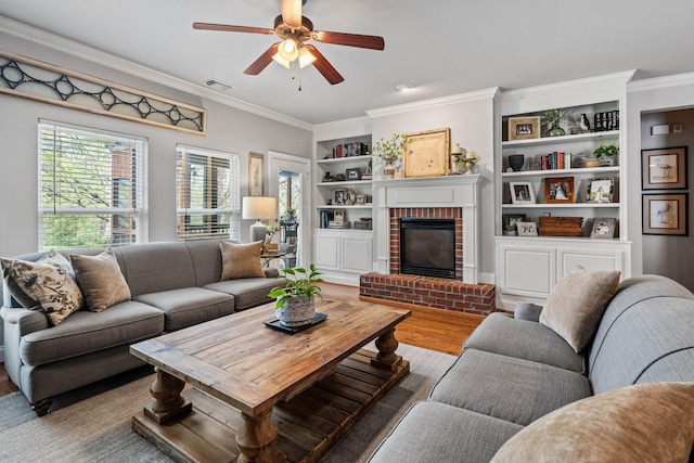 living area featuring ceiling fan, built in features, a brick fireplace, wood finished floors, and ornamental molding