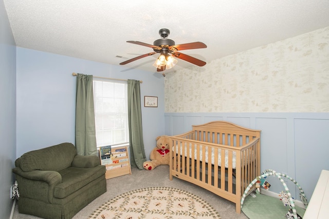 bedroom featuring carpet floors, visible vents, wainscoting, a nursery area, and a ceiling fan