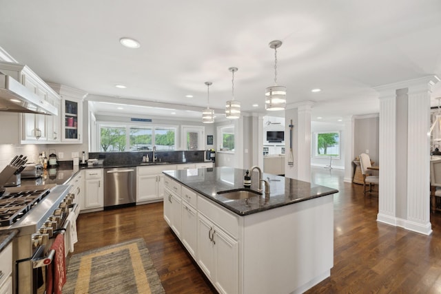 kitchen with appliances with stainless steel finishes, a sink, an island with sink, ornate columns, and glass insert cabinets