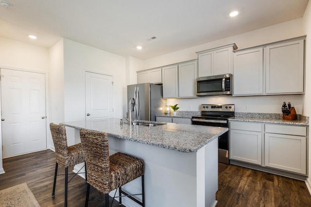 kitchen featuring a kitchen island with sink, a breakfast bar, dark wood finished floors, appliances with stainless steel finishes, and gray cabinetry