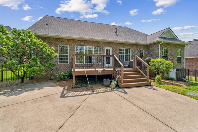 rear view of property featuring fence, a shingled roof, brick siding, a deck, and a patio