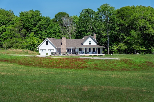 farmhouse inspired home featuring a front lawn, a garage, a porch, a chimney, and a wooded view