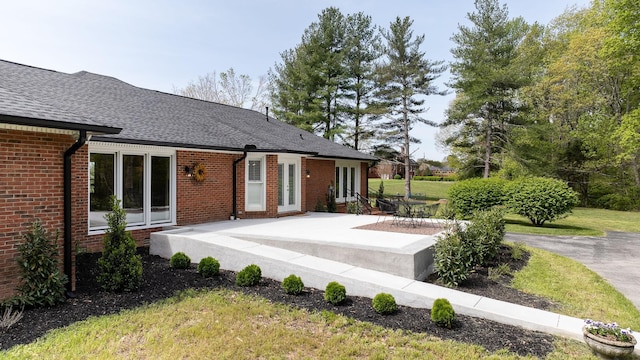 rear view of property with brick siding, a patio, a shingled roof, and a lawn