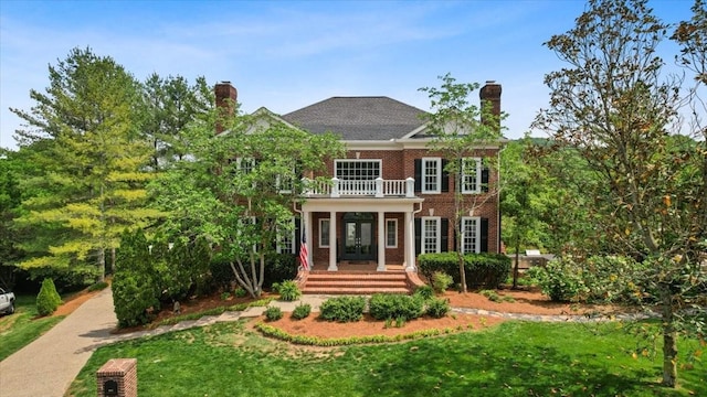 view of front of house with a front lawn, brick siding, a chimney, a balcony, and french doors
