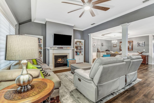 living area with decorative columns, a fireplace with flush hearth, wood finished floors, ceiling fan with notable chandelier, and ornamental molding