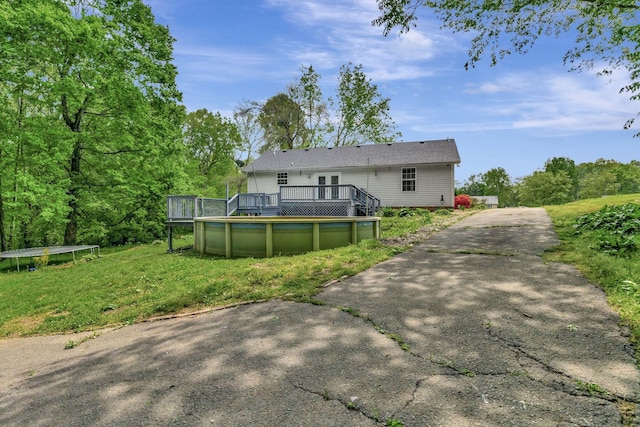 rear view of house with a trampoline, a lawn, a wooden deck, an outdoor pool, and aphalt driveway