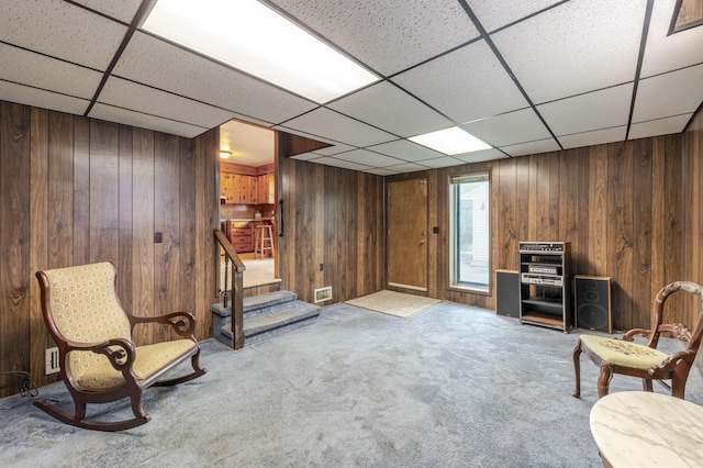 living area with carpet flooring, stairs, a drop ceiling, and wood walls