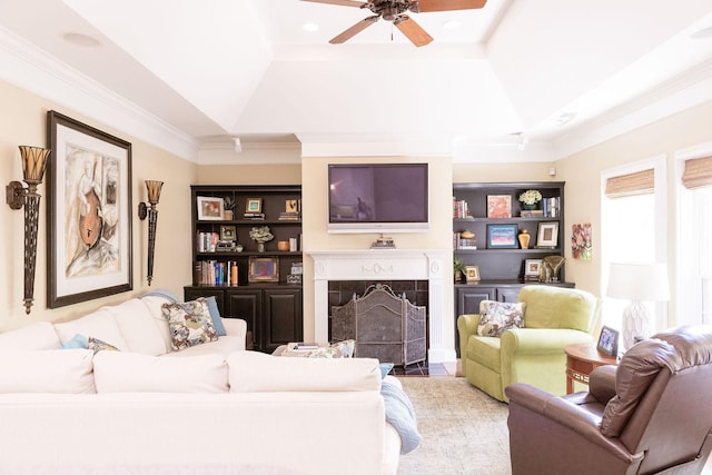 living room with ceiling fan, a premium fireplace, built in features, a tray ceiling, and ornamental molding