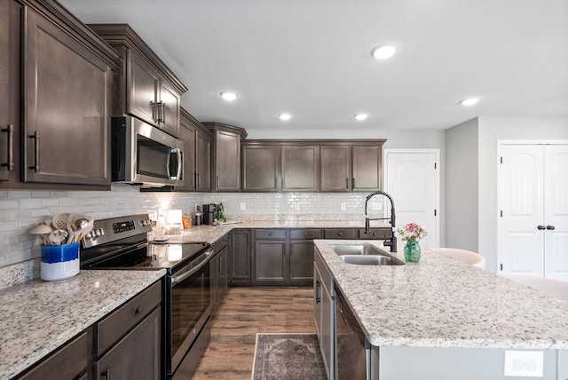 kitchen with an island with sink, dark brown cabinets, a sink, stainless steel appliances, and dark wood-type flooring
