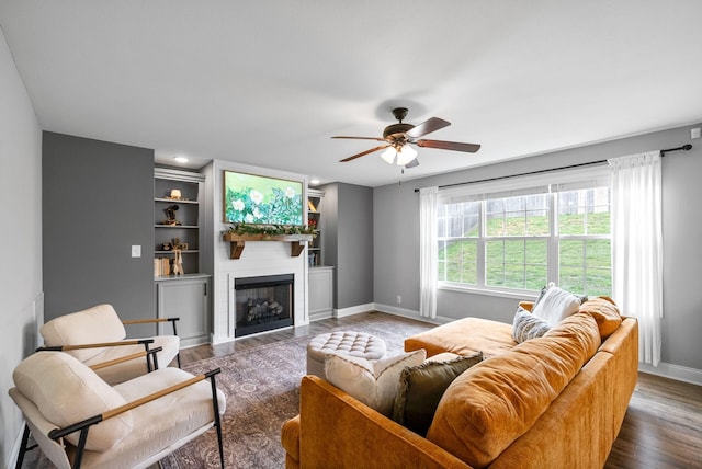 living room with a ceiling fan, a large fireplace, built in shelves, wood finished floors, and baseboards