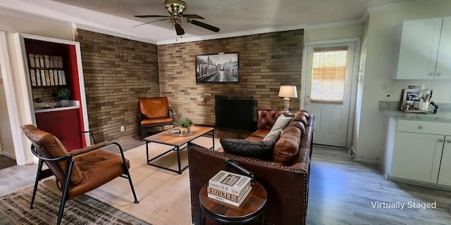 living area with a ceiling fan, brick wall, crown molding, light wood-type flooring, and baseboards