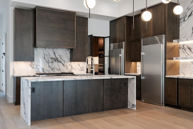 kitchen with dark brown cabinets, appliances with stainless steel finishes, light wood-style floors, decorative backsplash, and a center island with sink