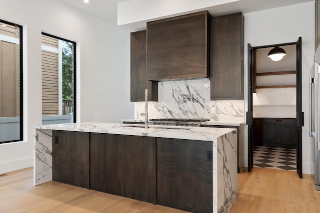 kitchen with backsplash, a center island with sink, dark brown cabinets, light stone counters, and light wood-style flooring