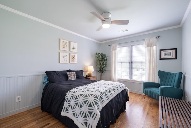 bedroom with crown molding, wainscoting, a ceiling fan, wood finished floors, and visible vents