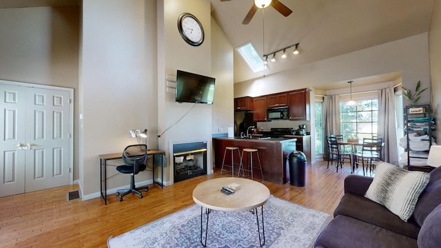 living room with ceiling fan, a skylight, rail lighting, high vaulted ceiling, and light wood finished floors