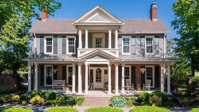 greek revival house featuring a porch, a shingled roof, brick siding, and a chimney