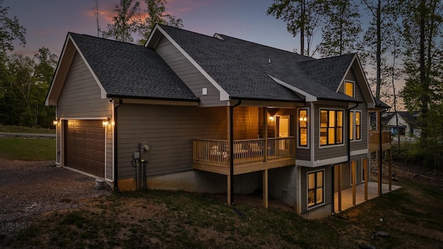 back of property at dusk featuring roof with shingles, an attached garage, a lawn, and driveway