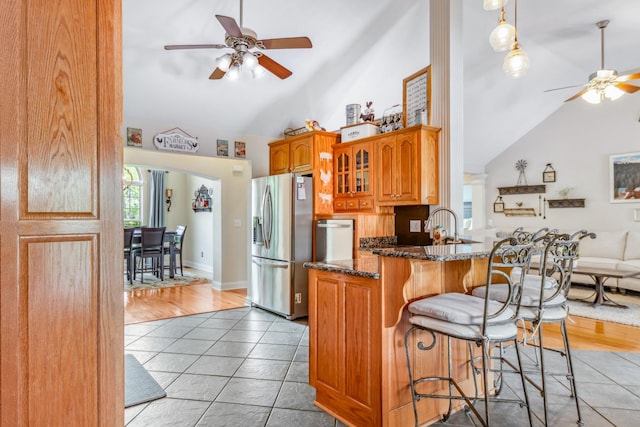 kitchen featuring glass insert cabinets, a ceiling fan, dark stone countertops, stainless steel fridge, and a breakfast bar