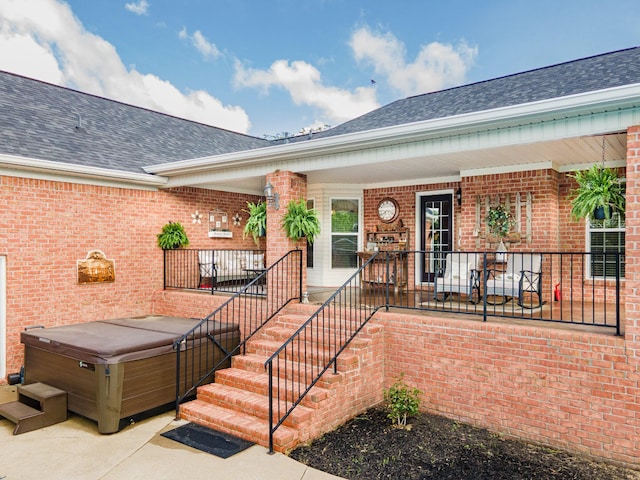 view of exterior entry featuring roof with shingles, a hot tub, and brick siding