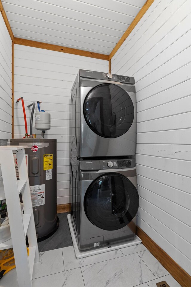 washroom featuring wooden walls, stacked washer / drying machine, laundry area, marble finish floor, and water heater