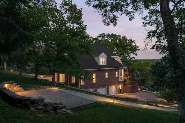 back of property at dusk featuring a lawn, a garage, driveway, and brick siding