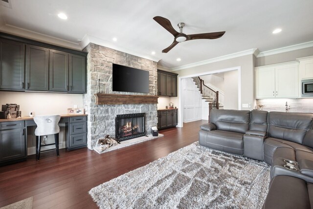 living area featuring crown molding, stairs, a stone fireplace, dark wood-type flooring, and a ceiling fan