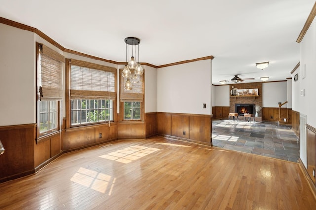 unfurnished dining area with a brick fireplace, a ceiling fan, wood-type flooring, and a wainscoted wall